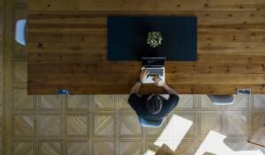 Eine Person arbeitet an einem Laptop an einem großen Holztisch in einem modernen Büro mit hellem Parkettboden und zeigt das Flexdesk Konzept mit flexiblen Arbeitsplätzen.