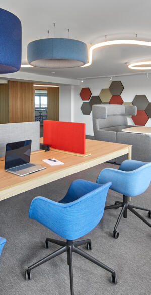 Helles Büro mit großen Akustikpaneelen, flexiblen Sitzbereichen, blauem Mobiliar und einem geteilten Arbeitstisch, das die zonierte Gestaltung für hybrides Arbeiten zeigt.
