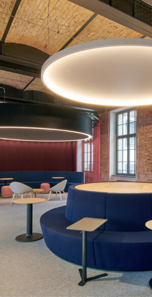 Offene Lounge mit runden Sitzinseln, kleinen Arbeitstischen und warmem Licht, die eine vertrauensvolle, flexible Umgebung für Hybrides Arbeiten und teamorientierte Zusammenarbeit zeigt.