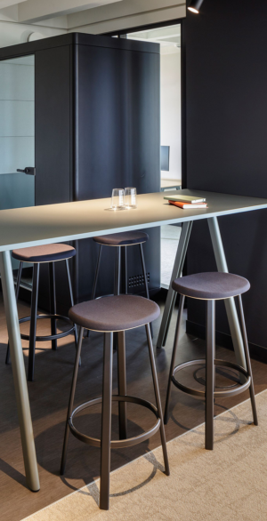 Hoher Stehtisch mit vier Hockern und zwei Gläsern vor einer modernen Meetingbox, die eine flexible Umgebung für hybrides Arbeiten zeigt.
