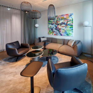 Fotografie eines Lounge-Bereichs einer modernen Bürofläche, der für eine gute Workplace Experience steht. Die Lounge ist mit diversen Sitzmöbel, wie Sofas und Sessel, kleinen Tische, einem akustischen Vorhang und Leuchten gestaltet. Die modernen und gedeckten Farben vermitteln Seriosität und Modernität und verleihen eine Wohlfühlatmosphäre, die für eine Workplace Experience wichtig ist. Ein modernes Gemälde mit bunten Farben auf einer Leinwand rundet die Szenerie ab.